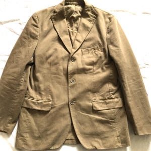 May include: A tan blazer with a notched lapel, three-button closure, and two front pockets. The jacket is made of a solid-colored fabric and has a tailored fit. The blazer is a classic piece of apparel.