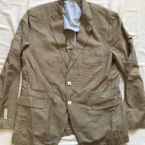 May include: A tan blazer with a subtle checkered pattern. The jacket features a notched lapel, two front pockets, and a two-button closure. The interior lining is a blue and white striped fabric. The sleeves have four buttons at the cuff.