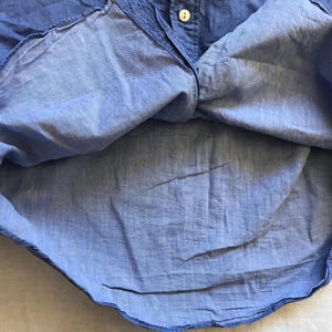 May include: Close-up of a light blue button-down shirt. The shirt is made of a lightweight fabric and features a classic collar and button closure. The fabric has a slightly wrinkled texture, suggesting it's made of linen or a similar material.