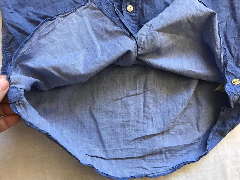 May include: Close-up of a light blue button-down shirt. The shirt is made of a lightweight fabric and features a classic collar and button closure. The fabric has a slightly wrinkled texture, suggesting it's made of linen or a similar material.