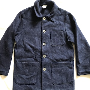 Puede incluir: Una chaqueta de trabajo azul marino con cierre de botones, dos bolsillos grandes en la parte inferior y un bolsillo más pequeño en el pecho. La chaqueta está hecha de una tela gruesa y texturizada.