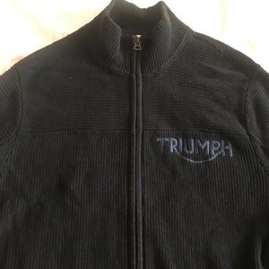Op de afbeelding: Zwarte trui met rits, geribbelde textuur en een blauw geborduurd Triumph-logo.