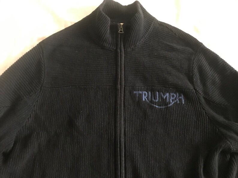 Op de afbeelding: Zwarte trui met rits, geribbelde textuur en een blauw geborduurd Triumph-logo.