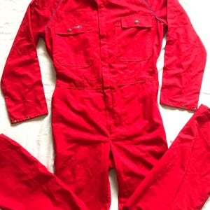 May include: Bright red jumpsuit with a button-up front, two chest pockets, and long sleeves. The jumpsuit has a collar and blue piping along the shoulders. The garment is made of a durable fabric.