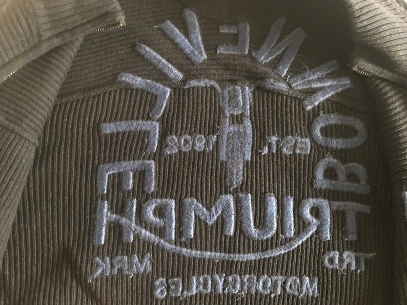 Op de afbeelding: Een zwart gebreid jack met een blauw geborduurd logo met de tekst "Triumph" en "Motocross" op de rug.