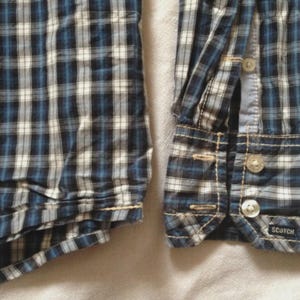 May include: Close-up of a blue, white, and black plaid shirt. The shirt features a buttoned cuff with two clear buttons and the brand name "SCOTCH" visible. The fabric is a woven cotton.