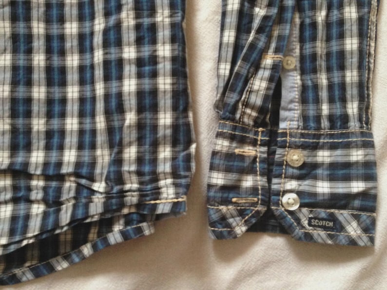May include: Close-up of a blue, white, and black plaid shirt. The shirt features a buttoned cuff with two clear buttons and the brand name "SCOTCH" visible. The fabric is a woven cotton.