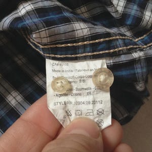 May include: Close-up of a blue, white, and black plaid shirt. The shirt's tag is visible, showing the style number and care instructions. The tag also includes the text "Made in India".