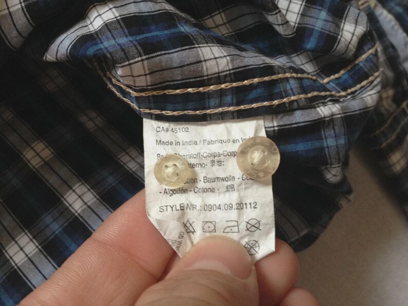 May include: Close-up of a blue, white, and black plaid shirt. The shirt's tag is visible, showing the style number and care instructions. The tag also includes the text "Made in India".