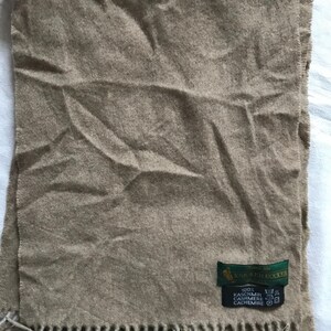 May include: A tan cashmere scarf with fringe. The scarf has a green label that reads "Knickerbocker" and "100% Cashmere Cashmere Cachemire".