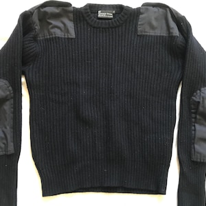 Może przedstawiać: Czarny sweter z prążkowanej dzianiny z wzmocnionymi naszywkami na ramionach i łokciach. Sweter ma okrągły dekolt i długie rękawy ze ściągaczami. Naszywki są wykonane z gładkiej, ciemnej tkaniny.