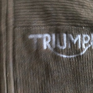 Op de afbeelding: Een close-up van een bruine gebreide stof met het woord "TRIUMPH" in wit geborduurd.