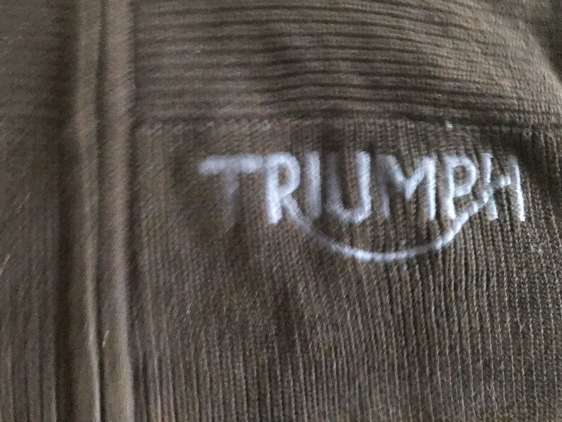 Op de afbeelding: Een close-up van een bruine gebreide stof met het woord "TRIUMPH" in wit geborduurd.
