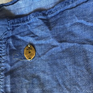 May include: Close-up of a blue denim shirt. The shirt features a small, oval, gold-coloured tag with the word "Scotch" and a palm tree design. The fabric is textured, showing stitching details.