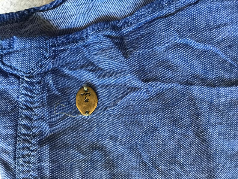 May include: Close-up of a blue denim shirt. The shirt features a small, oval, gold-coloured tag with the word "Scotch" and a palm tree design. The fabric is textured, showing stitching details.