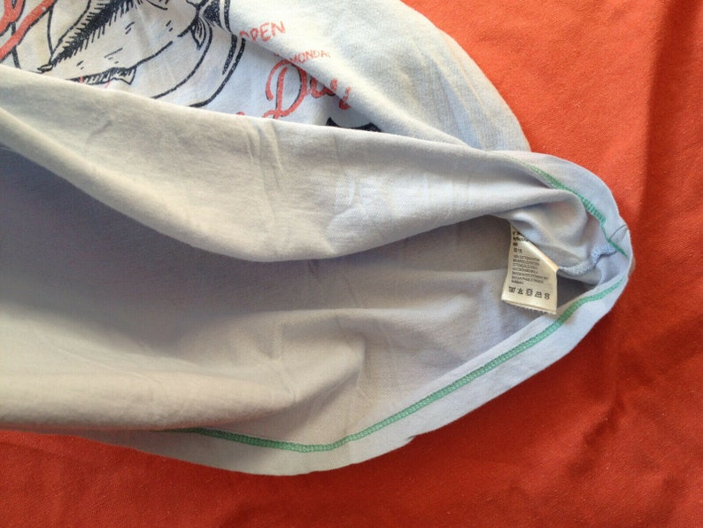 以下が含まれることがあります： 緑色の縁取りが施された薄い青色のTシャツ。Tシャツには魚のグラフィックプリントと「月曜日から金曜日まで営業」というテキストが書かれています。