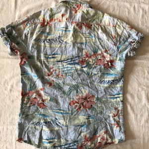 May include: A light blue short-sleeved button-down shirt with a tropical floral print. The print features red, pink, and green flowers, palm trees, and blue waves.