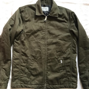 May include: Olive green jacket with a zip-up front, two front pockets, and a small pocket on the chest. The jacket has a collar and buttoned cuffs. The Carhartt logo is visible on the chest pocket.
