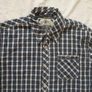 May include: A button-down shirt with a blue, white, and black plaid pattern. The shirt has a collared neckline, a chest pocket, and a button closure. The brand name "SCOTCH" is visible on a label near the collar.