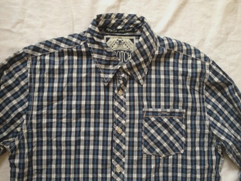 May include: A button-down shirt with a blue, white, and black plaid pattern. The shirt has a collared neckline, a chest pocket, and a button closure. The brand name "SCOTCH" is visible on a label near the collar.
