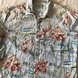 May include: A blue, short-sleeved button-down shirt with a tropical print featuring red, pink, and yellow flowers, green leaves, and blue waves. The shirt has a light blue background and a white collar.