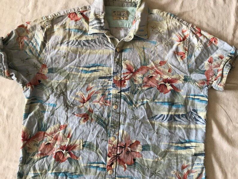 May include: A blue, short-sleeved button-down shirt with a tropical print featuring red, pink, and yellow flowers, green leaves, and blue waves. The shirt has a light blue background and a white collar.