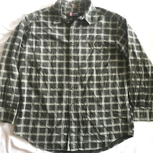 Puede incluir: Una camisa de manga larga con estampado de cuadros verdes y blancos. Tiene botones frontales y dos bolsillos en el pecho. Se aprecia el cuello y la etiqueta de la marca "Chaps". La tela parece suave y cómoda.