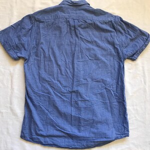 May include: A short-sleeved, blue button-down shirt. The shirt is made of a textured fabric and has a button-down collar. The back of the shirt is visible, showing the yoke and the button detail. The shirt is laid flat on a white surface.