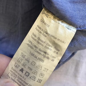May include: A close-up shot of a clothing label with text in multiple languages, including English, French, and Portuguese. The label indicates the item is made in India and is 100% cotton. Washing instructions are visible.