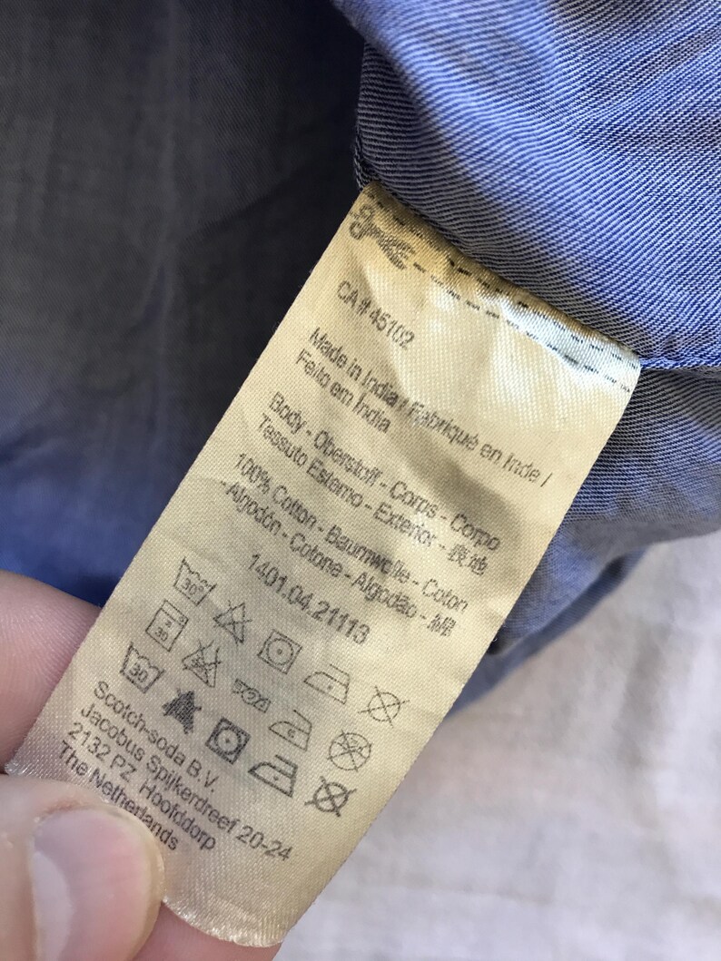May include: A close-up shot of a clothing label with text in multiple languages, including English, French, and Portuguese. The label indicates the item is made in India and is 100% cotton. Washing instructions are visible.