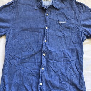 May include: A blue short-sleeved shirt with a button-down front, chest pocket, and collar. The shirt features white buttons and a small label on the pocket. The fabric is a textured cotton blend.