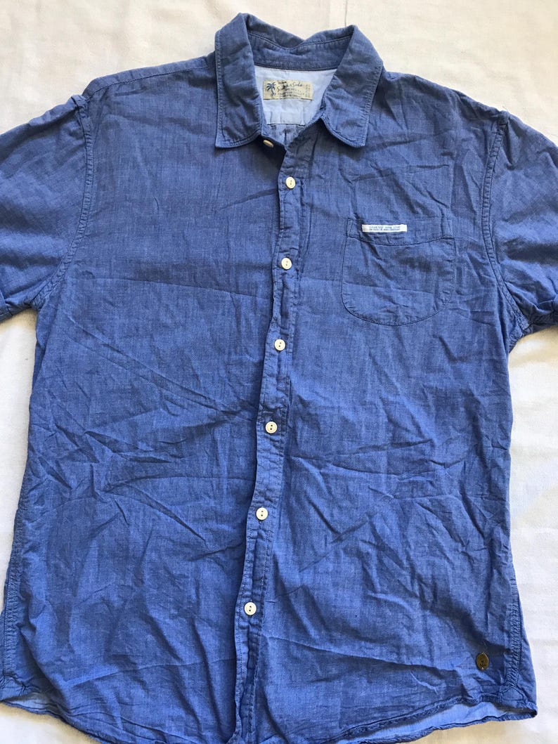 May include: A blue short-sleeved shirt with a button-down front, chest pocket, and collar. The shirt features white buttons and a small label on the pocket. The fabric is a textured cotton blend.