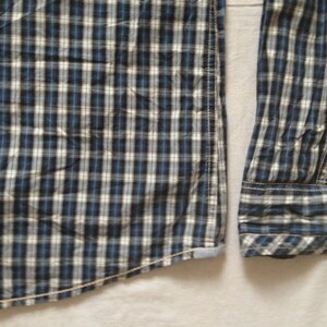 May include: Close-up of a blue, white, and black plaid button-down shirt. The long-sleeved shirt has a classic collar and cuffs. The fabric is a woven cotton, and the shirt is partially unbuttoned.