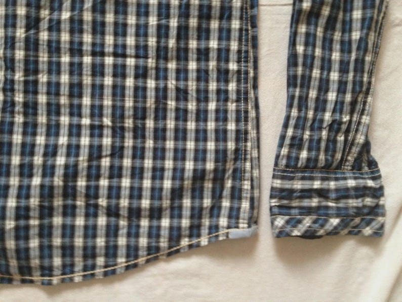 May include: Close-up of a blue, white, and black plaid button-down shirt. The long-sleeved shirt has a classic collar and cuffs. The fabric is a woven cotton, and the shirt is partially unbuttoned.