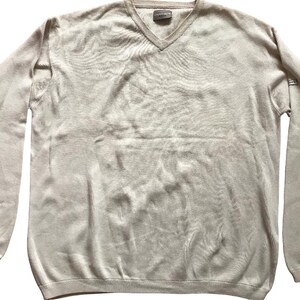 Puede incluir: Un suéter beige claro con cuello en V y mangas largas. El suéter tiene un dobladillo y puños acanalados. La tela parece ser un tejido suave. Una pequeña etiqueta es visible en el escote. El suéter está tendido sobre una superficie blanca.