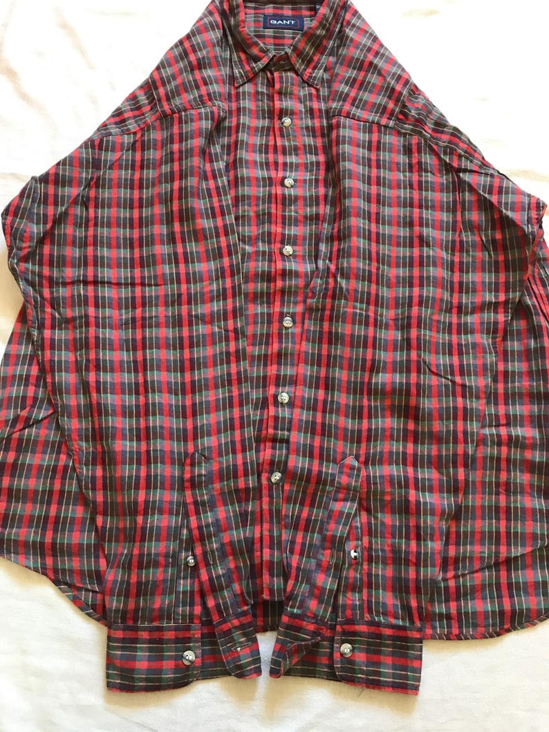 Puede incluir: Una camisa a cuadros roja, negra y verde con cuello y mangas largas. La camisa tiene botones plateados y la marca "GANT" es visible en el cuello. La camisa est&aacute; hecha de una tela ligera.