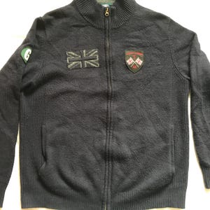 Puede incluir: Cárdigan con cremallera azul marino oscuro con textura acanalada. Cuenta con un parche de la Union Jack, un parche verde con una "G" blanca y un parche con escudo con banderas cruzadas. El cárdigan tiene cuello alto y dos bolsillos delanteros.