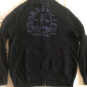 Op de afbeelding: Zwarte gebreide cardigan met rits en een blauw geborduurd logo met de tekst "Bonneville Triumph Motorcycles" en "Est. 1902."
