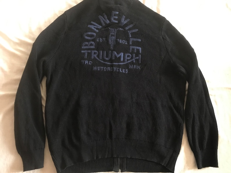 Op de afbeelding: Zwarte gebreide cardigan met rits en een blauw geborduurd logo met de tekst "Bonneville Triumph Motorcycles" en "Est. 1902."