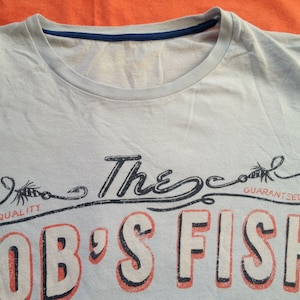以下が含まれることがあります： 薄い青色のTシャツに、釣り針のグラフィックプリントと「The Cool Guaranteed Bob's Fish」という赤い文字と黒い文字がプリントされています。