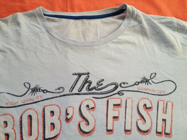 以下が含まれることがあります： 薄い青色のTシャツに、釣り針のグラフィックプリントと「The Cool Guaranteed Bob's Fish」という赤い文字と黒い文字がプリントされています。