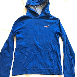 May include: A long-sleeve blue hooded sweatshirt featuring a front kangaroo pocket and a small red and white bird logo embroidered on the chest. The interior of the hood is lined with a white and navy blue striped fabric pattern.