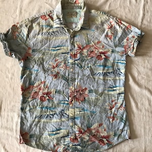 May include: A light blue short-sleeved button-down shirt with a tropical print featuring red, pink, and green flowers, palm trees, and blue waves.