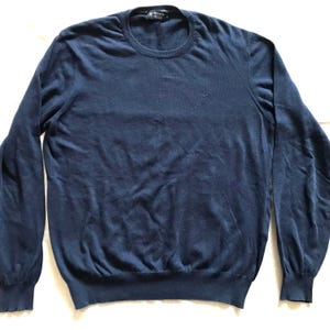 Puede incluir: Un jersey azul marino de cuello redondo con mangas largas. El jersey tiene un pequeño logotipo bordado en el pecho y cuello, puños y dobladillo acanalados. El nombre de la marca es visible en la etiqueta.