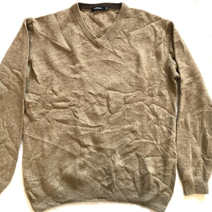 Puede incluir: Un suéter beige con cuello en V y mangas largas. El suéter tiene un dobladillo y puños acanalados. El escote es de un tono marrón más oscuro. La tela parece ser un material de punto suave.