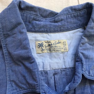 May include: A close-up of a blue denim shirt collar. The shirt features a label with the text "Scotch & Soda" and a palm tree logo. The fabric has a soft, textured appearance.