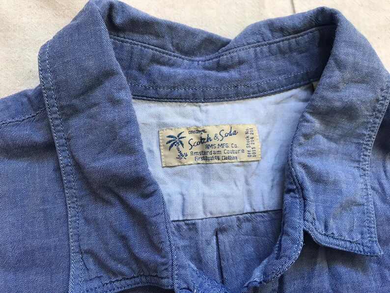 May include: A close-up of a blue denim shirt collar. The shirt features a label with the text "Scotch & Soda" and a palm tree logo. The fabric has a soft, textured appearance.