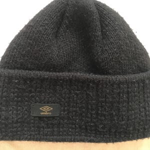 May include: A black knit beanie hat with a folded brim. The hat features a small, rectangular patch with the Umbro logo in gold. The beanie is made of a thick, textured material.