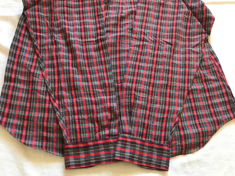 Puede incluir: Una camisa a cuadros roja, negra y verde. La camisa de manga larga tiene un cuello cl&aacute;sico. El patr&oacute;n de cuadros es un dise&ntilde;o tradicional, con los colores dispuestos en una cuadr&iacute;cula. Los pu&ntilde;os tambi&eacute;n son a cuadros.