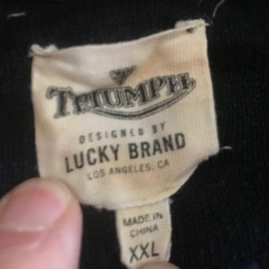 Op de afbeelding: Een wit label met zwarte tekst dat "Triumph Designed by Lucky Brand Los Angeles, CA Made in China XXL" zegt.